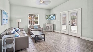 A spacious living room with a ceiling fan, a sofa set, a coffee table, a TV mounted on the wall, and a sliding glass door leading outside.