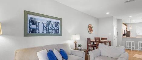 A living room with a beige sofa, two armchairs, a dining table with chairs, a clock, and a kitchen area in the background.