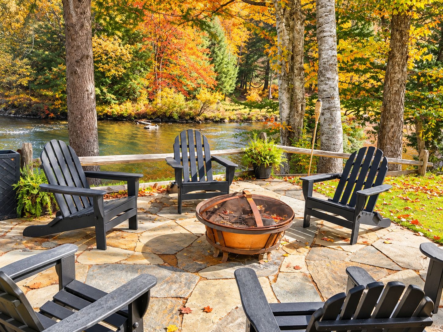 A patio with Adirondack chairs, a fire pit, and a view of a river and autumn foliage.