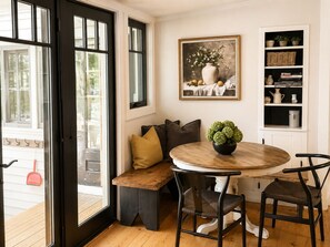 A dining area with a round wooden table, two chairs, a bench with cushions, a painting above the bench, and a shelf with decorative items.