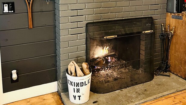 Una chimenea moderna con un televisor encima, un revestimiento decorativo y un cubo etiquetado "Kindle in VT" al lado de la chimenea.