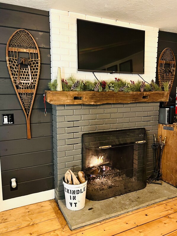 A modern fireplace with a TV above it, a decorative mantelpiece, and a bucket labeled "Kindle in VT" beside the fireplace.