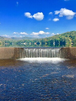 A waterfall over a concrete barrier with a lake and forested hills in the background.
