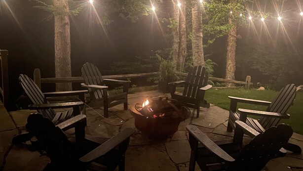 Un espacio al aire libre con sillas Adirondack y un brasero, rodeado de árboles y luces colgantes.