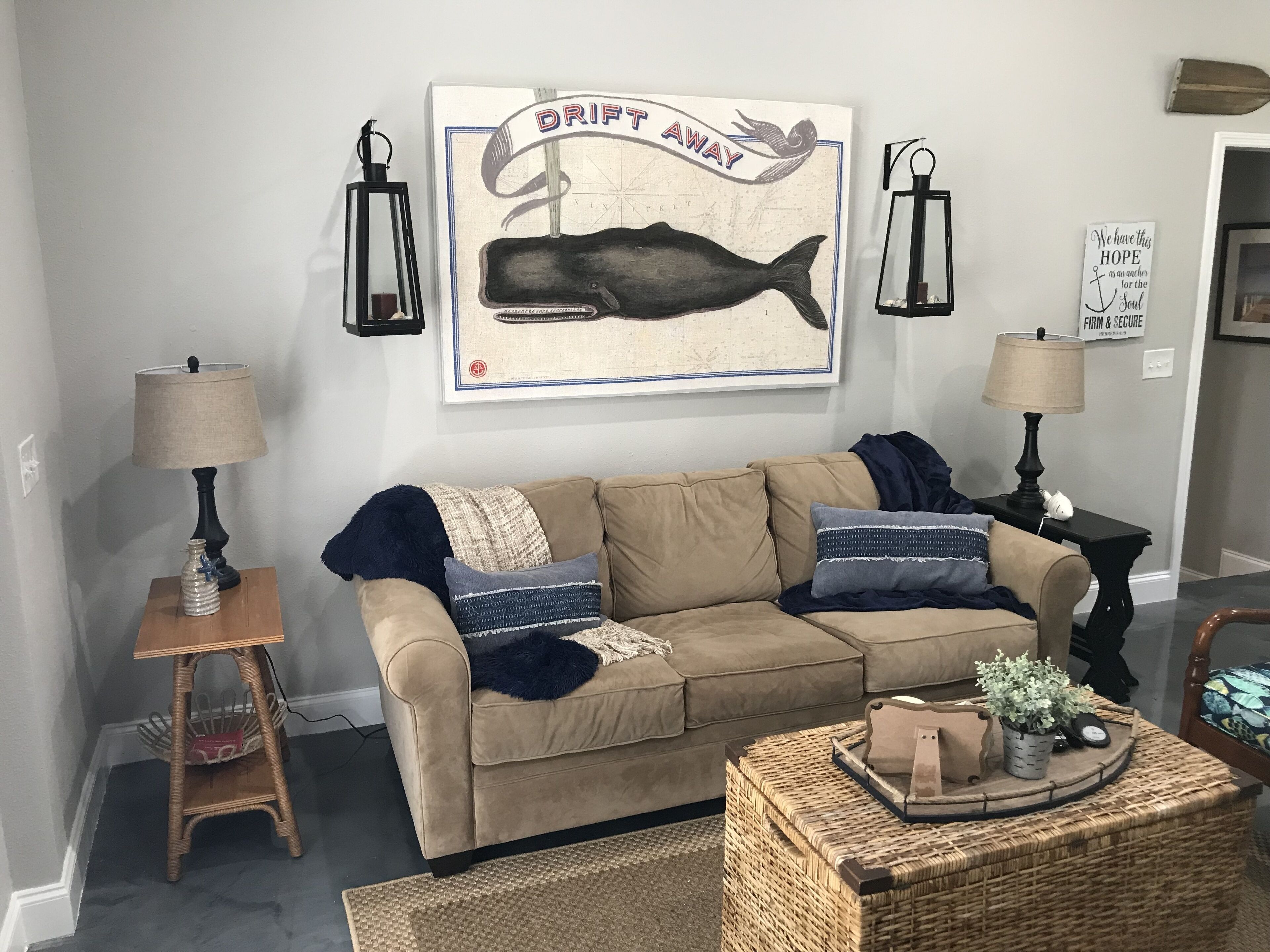 Una sala de estar con un sofá color beige, dos mesas auxiliares con lámparas, una obra de arte enmarcada sobre el sofá y una mesa de centro de mimbre con objetos decorativos.