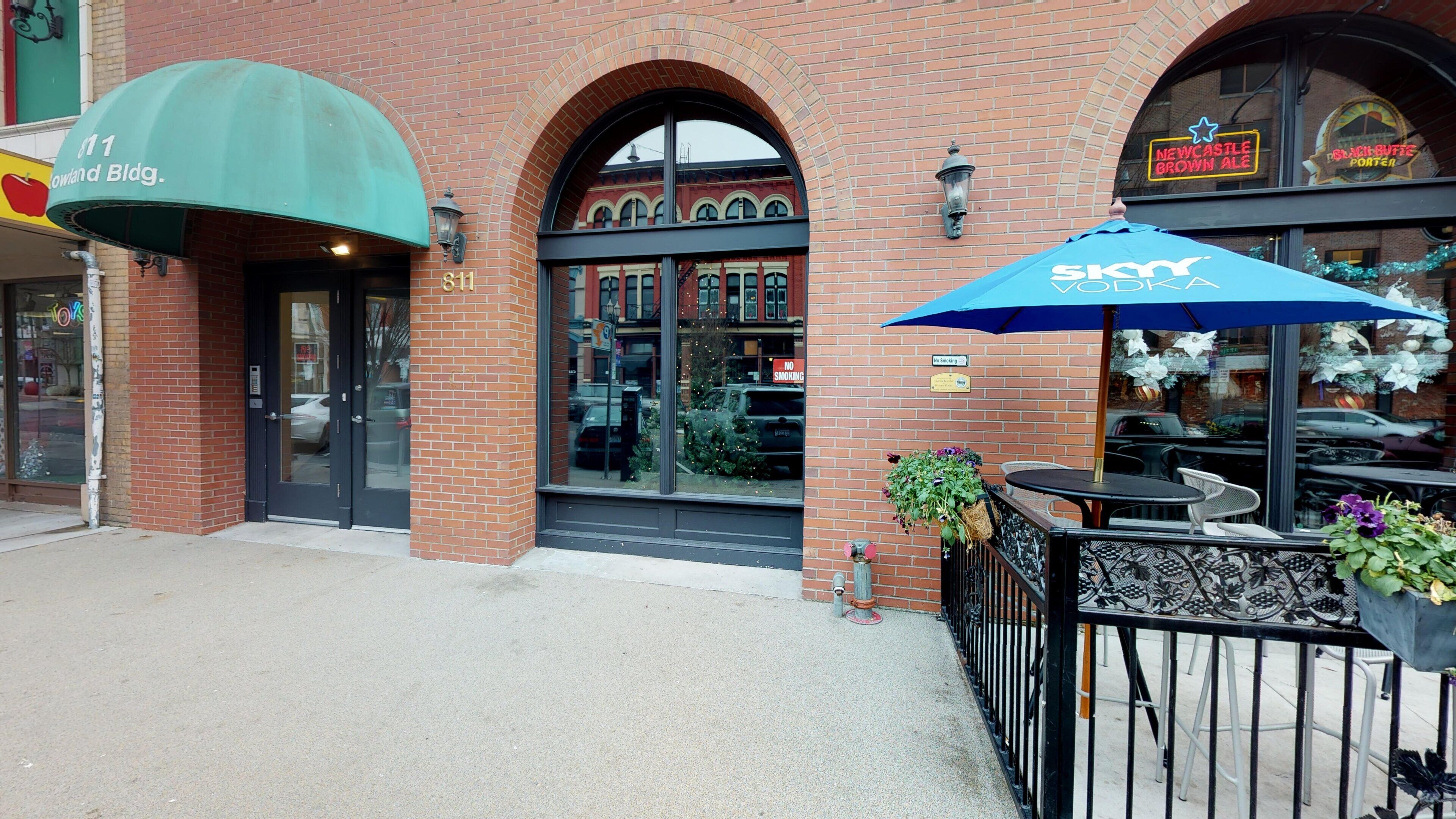 A brick building with a green awning, a storefront with a 'No Smoking' sign, and outdoor seating with a blue umbrella.