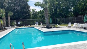 A rectangular swimming pool with a tiled edge, surrounded by a brick deck. There are lounge chairs and a table with umbrellas, and a fence with signs indicating no diving and no lifeguard on duty.