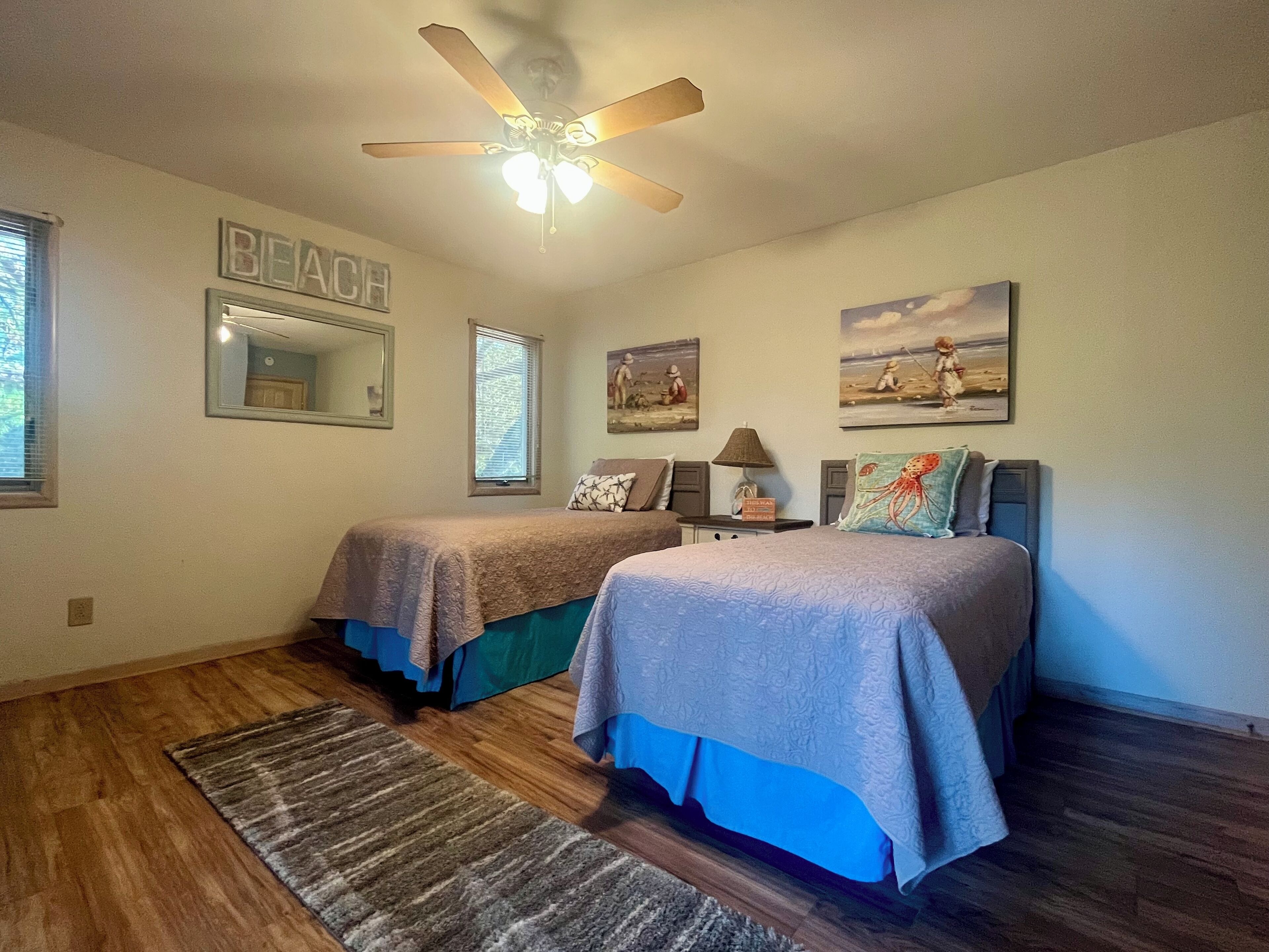 A bedroom with two beds, a ceiling fan, a beach-themed wall, and a rug.