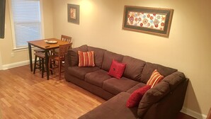 Una sala de estar con un sofá seccional marrón, dos almohadas rojas, una mesa de comedor de madera con sillas y una obra de arte enmarcada en la pared.