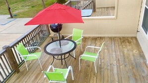 Una terraza de madera con una mesa redonda de vidrio y cuatro sillas verdes, un paraguas rojo y una parrilla negra.