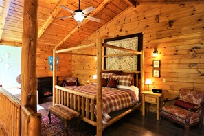 Un dormitorio de cabaña de madera con una cama con dosel, una chimenea, un ventilador de techo y una mesita de noche con lámpara.
