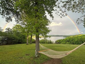A hammock tied to a tree in a grassy area with a lake and a rainbow in the background.