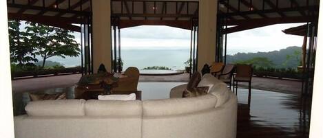 A spacious living area with a large sofa, a wooden table with a bowl of fruit, and a view of the ocean through open spaces.