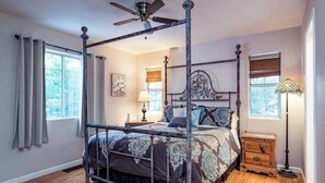 A four-poster bed with a ceiling fan, a bedside table, a lamp, and a window with curtains.