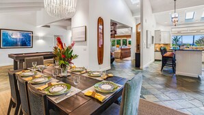 A spacious dining area with a long table set for six, a modern chandelier, and a view of the kitchen and living area. - Stunning Gated Home, Private Pool & Spa (Lahaina)