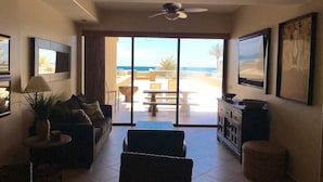 A living room with a sofa, armchair, and a coffee table. There is a TV mounted on the wall, and a ceiling fan above. The room has a sliding glass door leading to an outdoor patio with a table and chairs. - In The Middle Of All, Ocean Front Ground Floor 2BD. Steps from the beach Phase 1 (Puerto penasco)