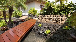 A wooden deck leading to a building with a stone wall featuring statues. - Ocean View Home (Kailua-Kona)