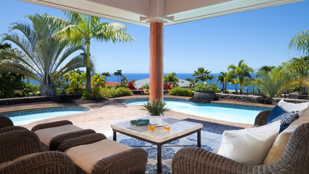 A poolside area with wicker furniture, a table with drinks, and a view of the ocean. - Ocean View Home (Kailua-Kona)
