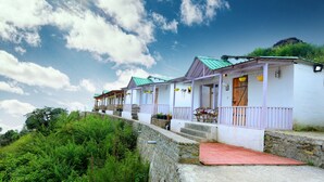 A row of white buildings with green roofs and purple railings, situated on a hillside with a stone retaining wall. - Banlekhi (Dhari)