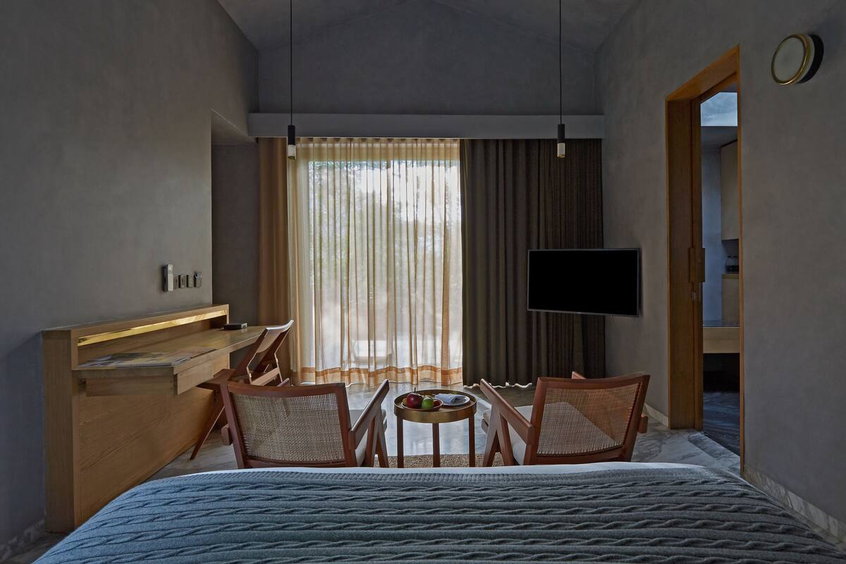 A hotel room with a bed, a desk, two chairs, a small table with fruits, a TV, and a window with curtains.
