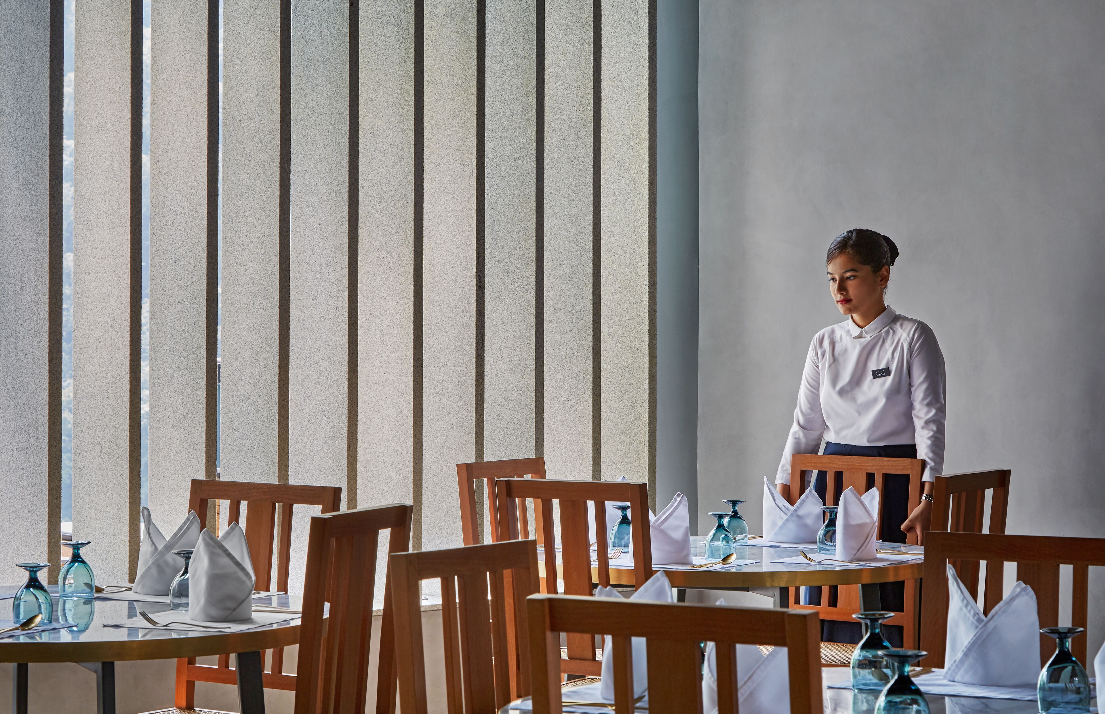 Een restaurant met houten stoelen en tafels, een bediende in uniform, en een groot raam met verticale jaloezieën.
