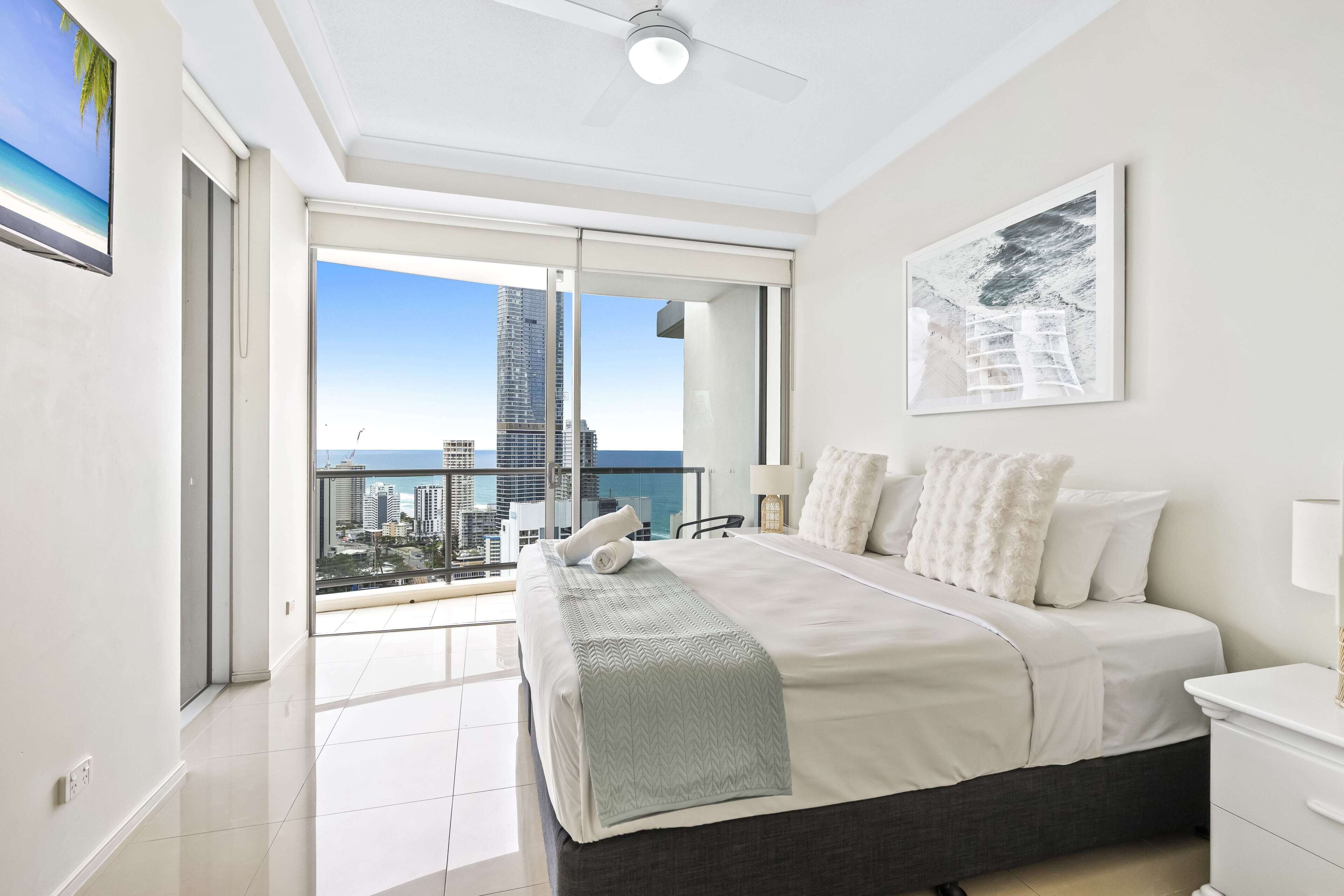 Ein modernes Schlafzimmer mit einem großen Bett, einem Balkon und Blick auf die Stadt sowie einem Deckenventilator.