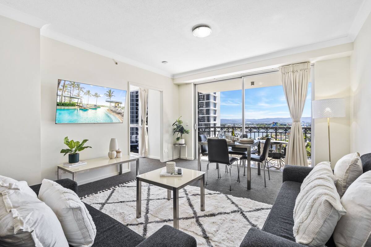 A modern living room with a large TV, a sofa, a coffee table, and a balcony with a view.