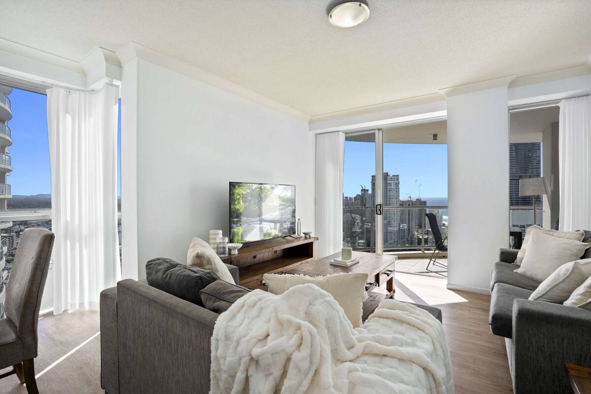 A modern living room with a flat-screen TV, grey sofas, a white throw blanket, and a balcony with a city view.