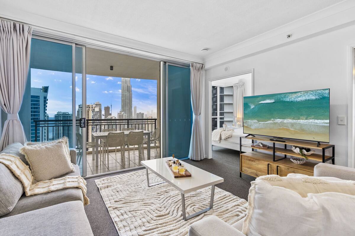 A modern living room with a large TV, a sofa, a coffee table, and a view of the city skyline through the balcony doors.