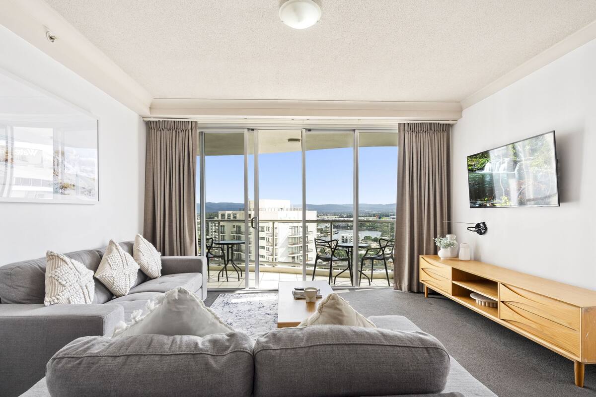 A modern living room with a grey sofa, a wooden TV stand, and a balcony with a city view.
