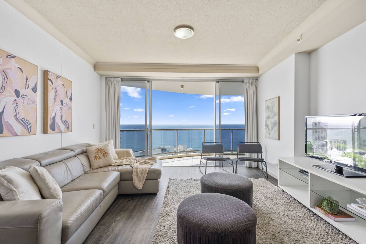 A modern living room with a sofa, ottoman, and TV stand, a balcony with a view of the sea, and artwork on the walls.