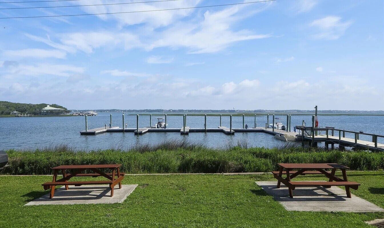 A lakeside picnic area with two wooden picnic tables, a dock extending into the water, and a grassy foreground.