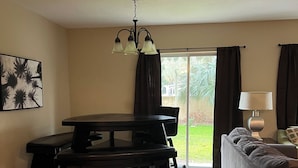 Un salon spacieux doté d’un plancher en bois, d’une table à manger avec des chaises, d’un canapé, d’une lampe et d’une grande fenêtre agrémentée de rideaux.