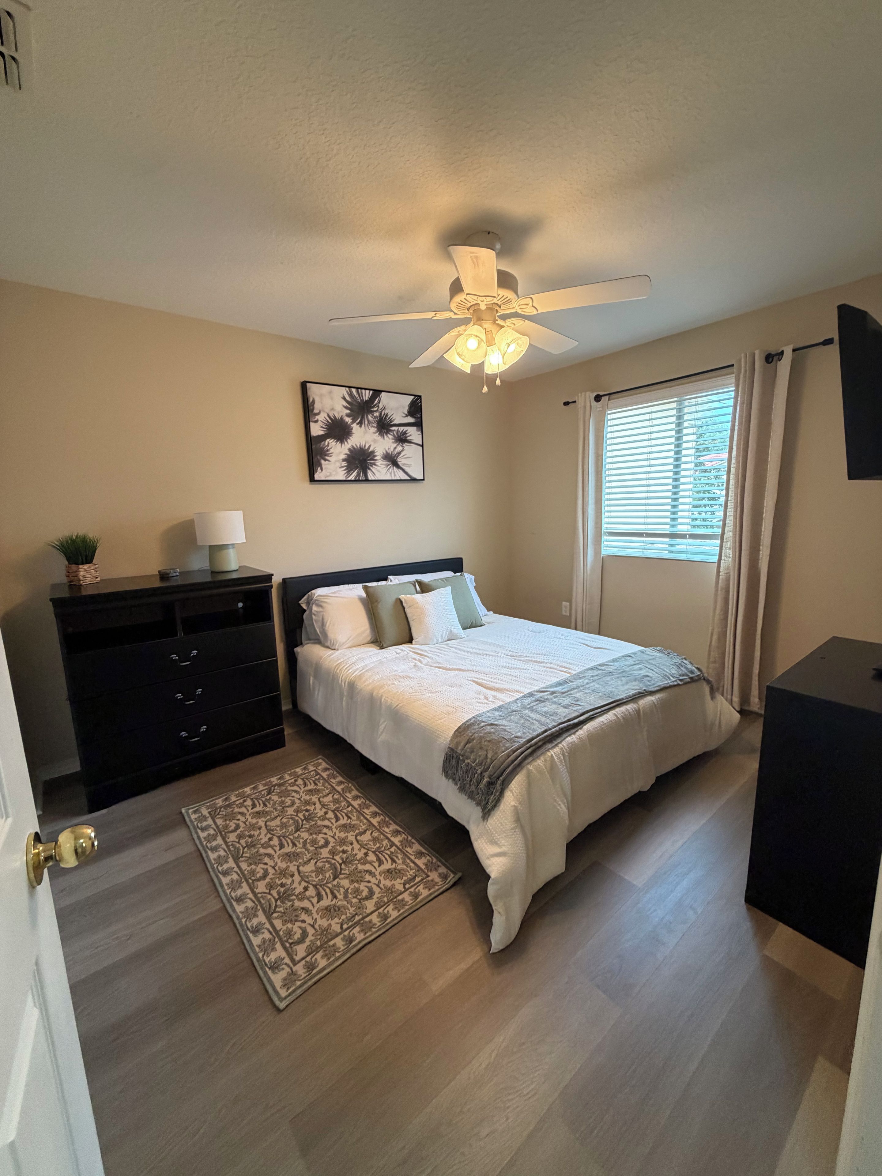 A bedroom with a bed, a dresser, a ceiling fan, a window with curtains, and a rug.