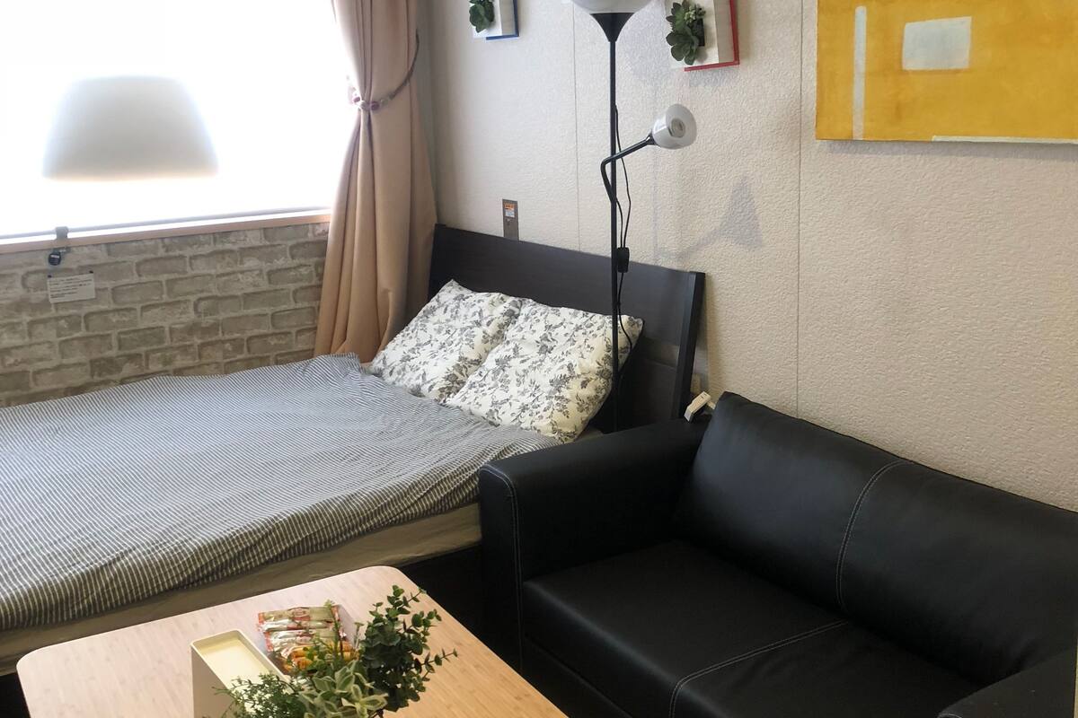 A room with a bed, a black leather sofa, a wooden table with plants, a floor lamp, and a wall-mounted air conditioner.