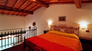Un dormitorio con techo de madera, cama con colcha roja y naranja, cabecera de metal, dos lámparas de noche y un cuadro pequeño en la pared.