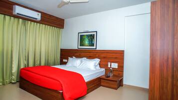 Habitación de hotel con cabecera de madera, cama con sábanas blancas y manta roja, mesita de noche con lámpara, equipo de aire acondicionado, cuadro en la pared y puerta con panel de madera.