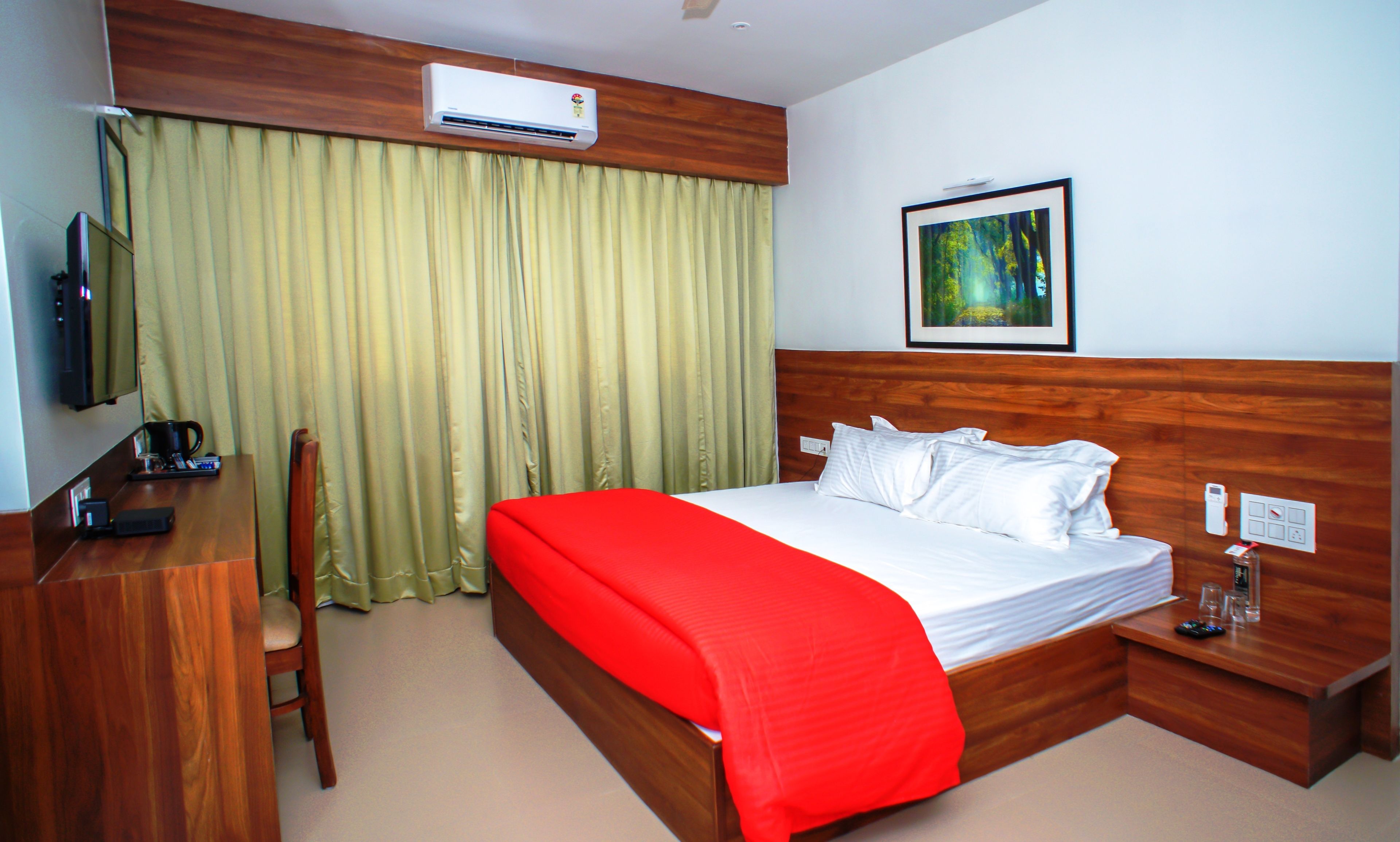 Habitación de hotel con cabecera de madera, cama con ropa de cama blanca y detalles en rojo, escritorio con cafetera, televisor y una imagen de un bosque.