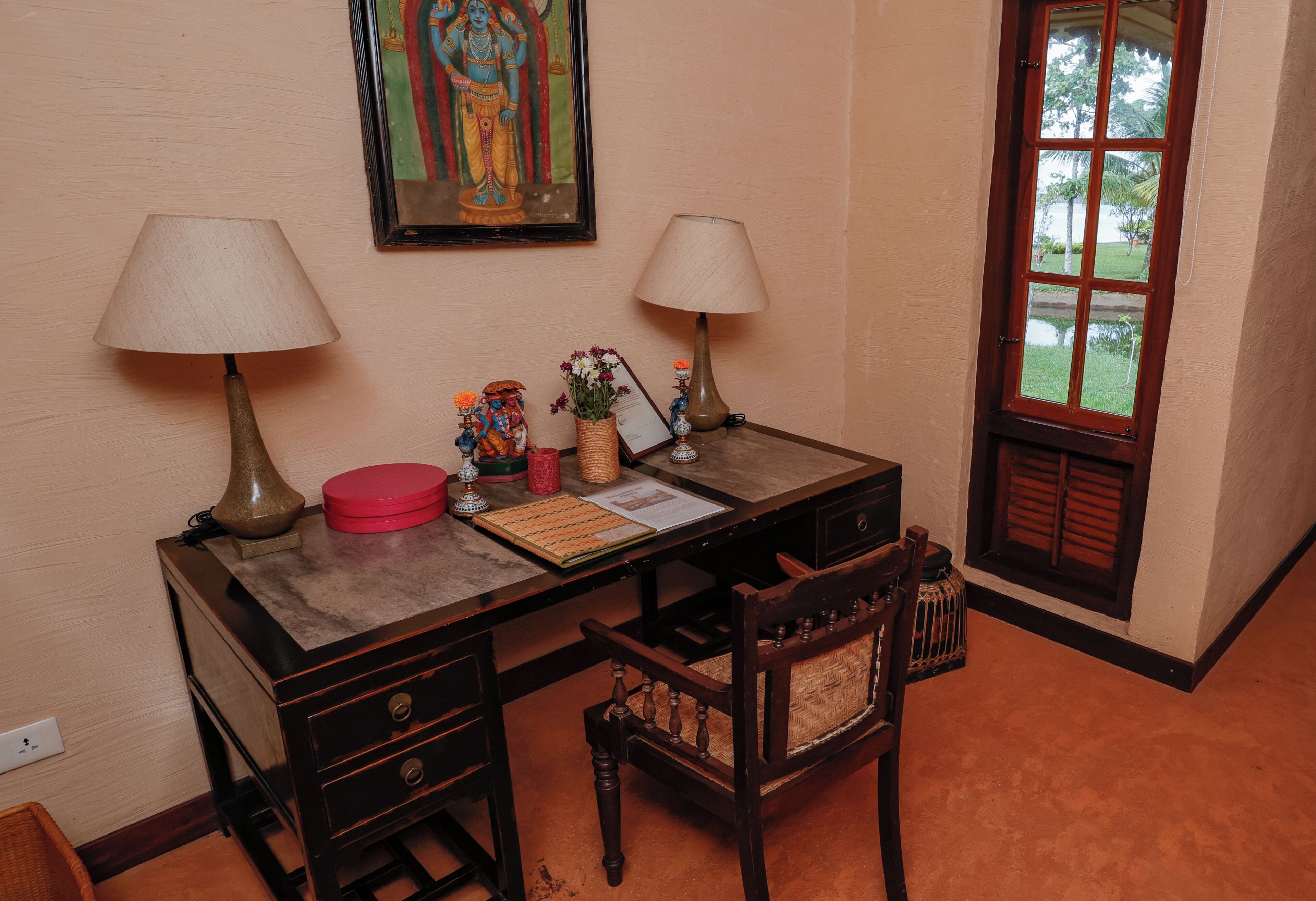 Una habitación con un escritorio, dos lámparas, una silla, una pequeña estatua, un jarrón con flores y un cuadro en la pared.