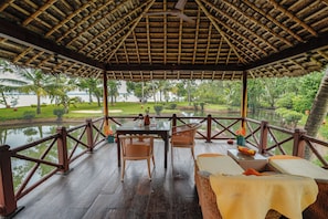 A covered outdoor seating area with a table and chairs, overlooking a body of water and greenery. - Ameya (Cherthala)