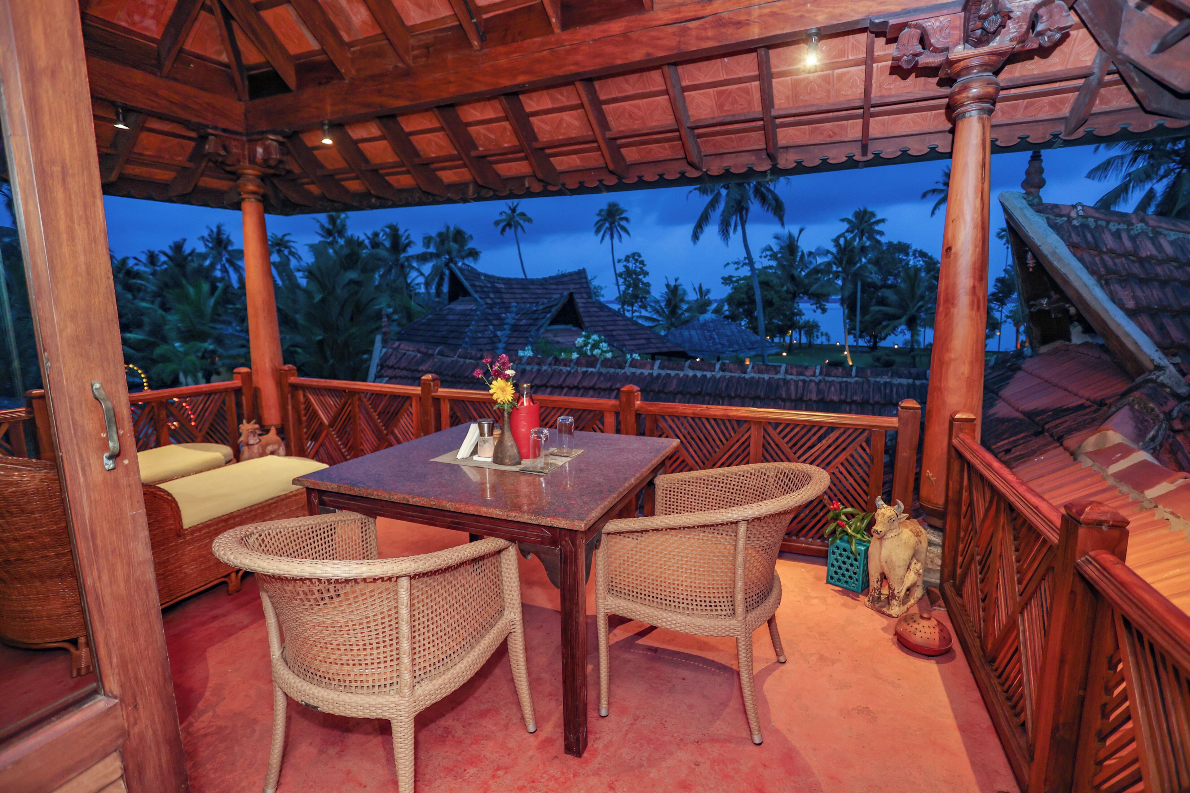 An outdoor seating area with a wooden table and wicker chairs, overlooking a tropical landscape with palm trees and traditional architecture.