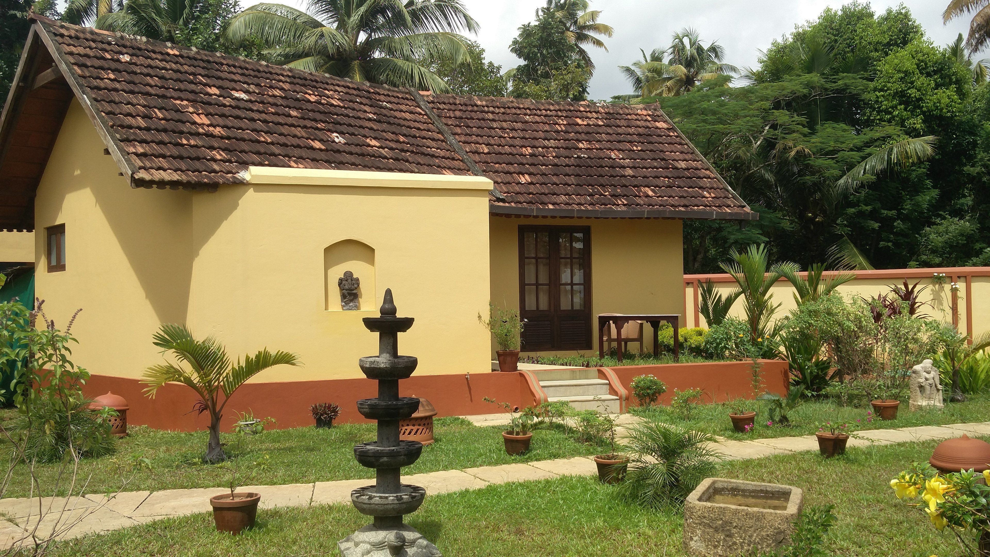 A small, yellow building with a tiled roof, surrounded by a garden with potted plants and a stone fountain.
