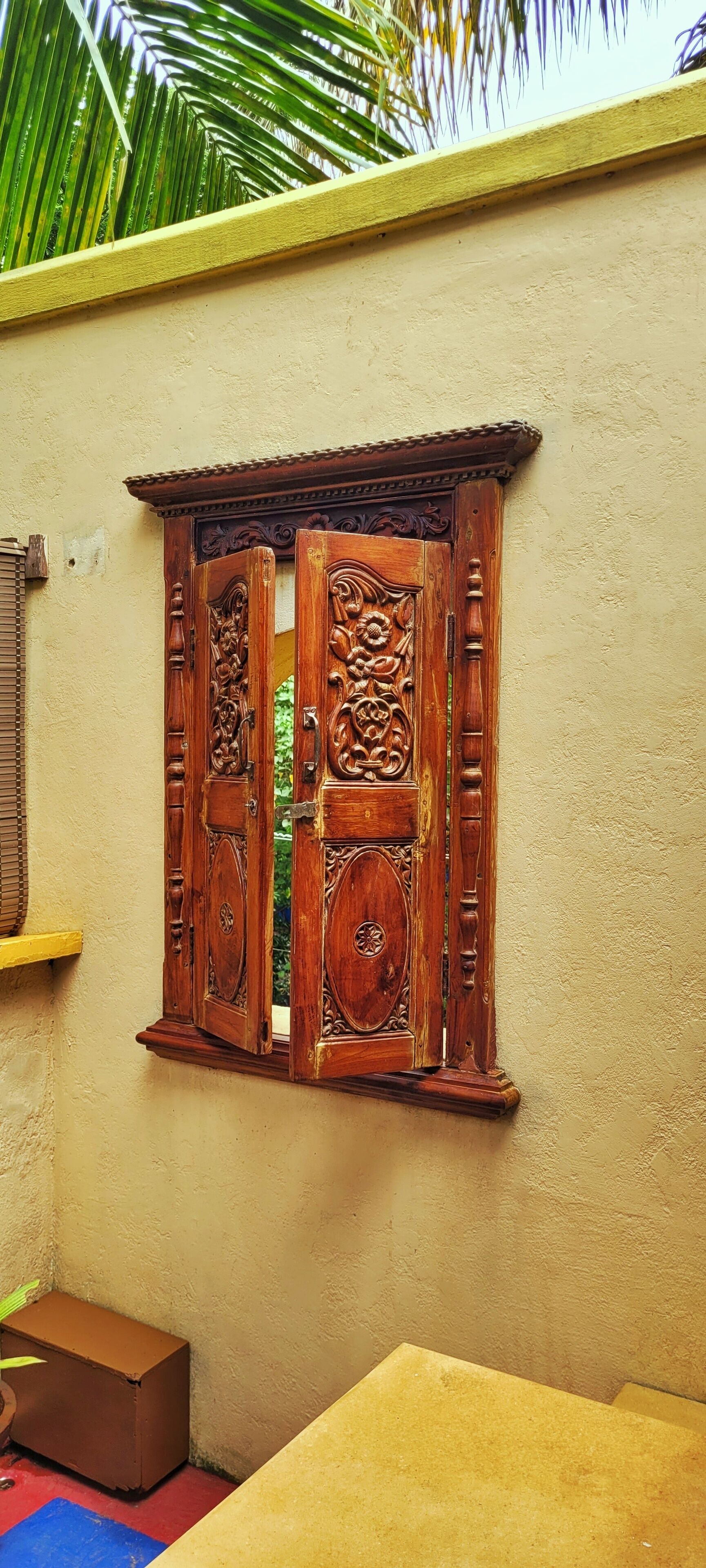 A wooden window with intricate carvings, set in a beige wall with a palm tree in the background.
