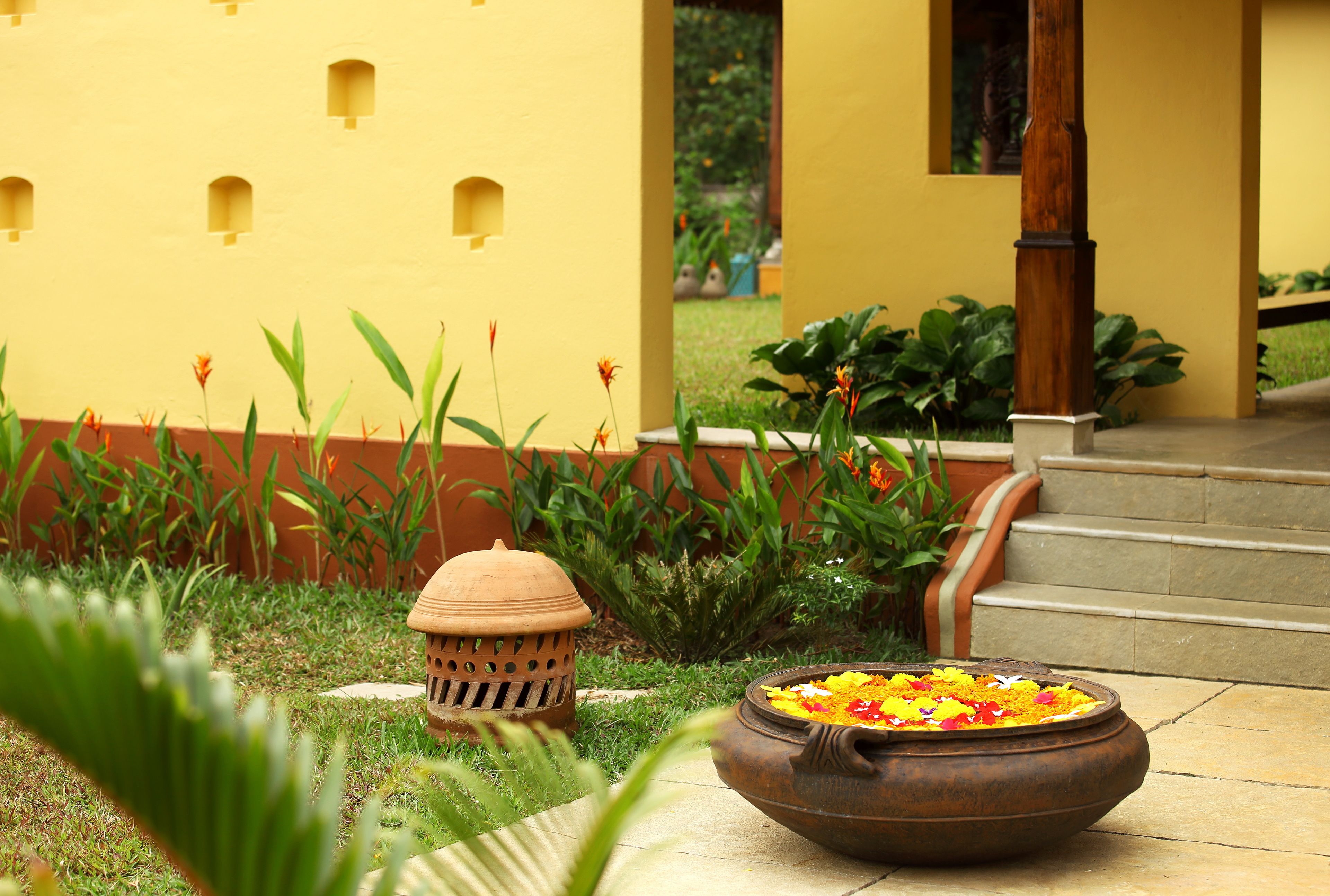 A courtyard with a large clay pot filled with flowers, a traditional lamp, and a yellow wall with small windows.