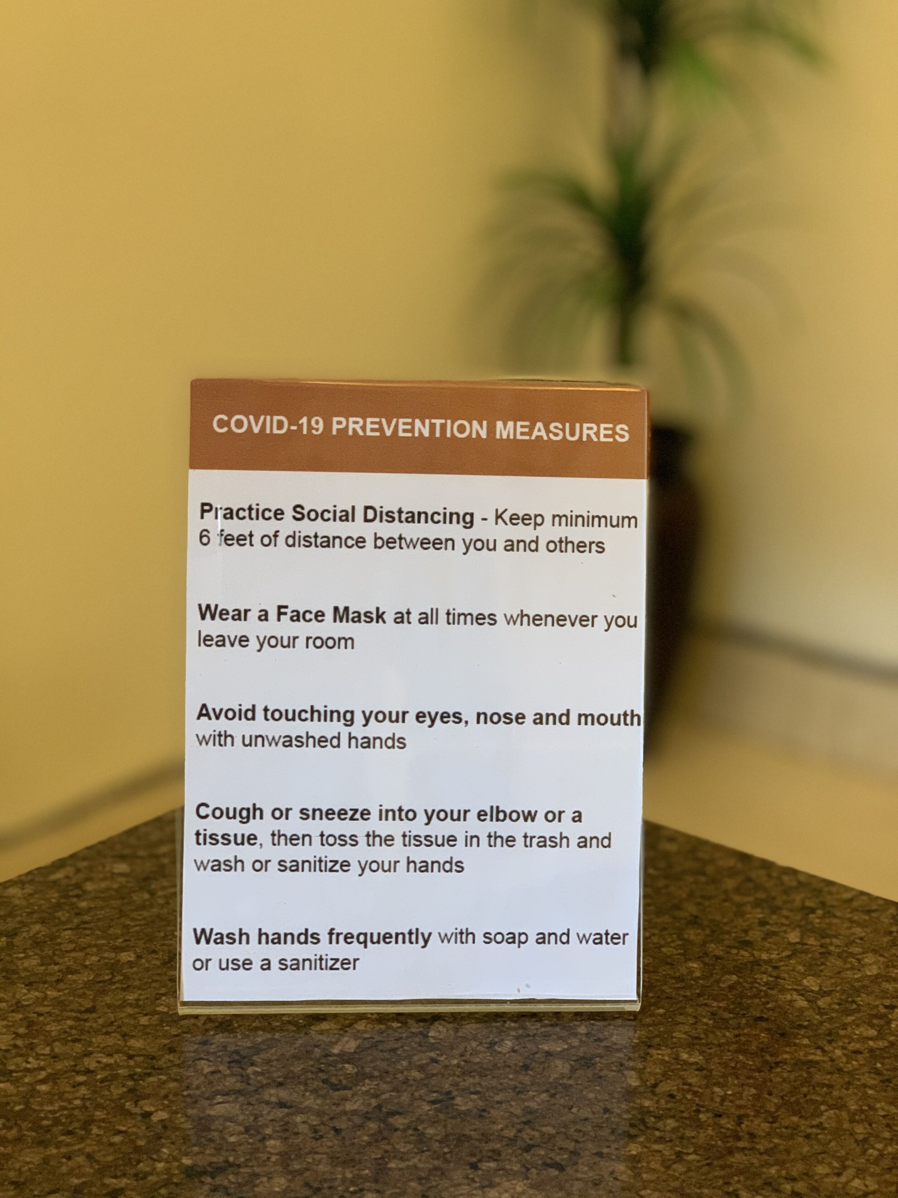 A sign with COVID-19 prevention measures including social distancing, wearing a face mask, avoiding touching face, and washing hands.
