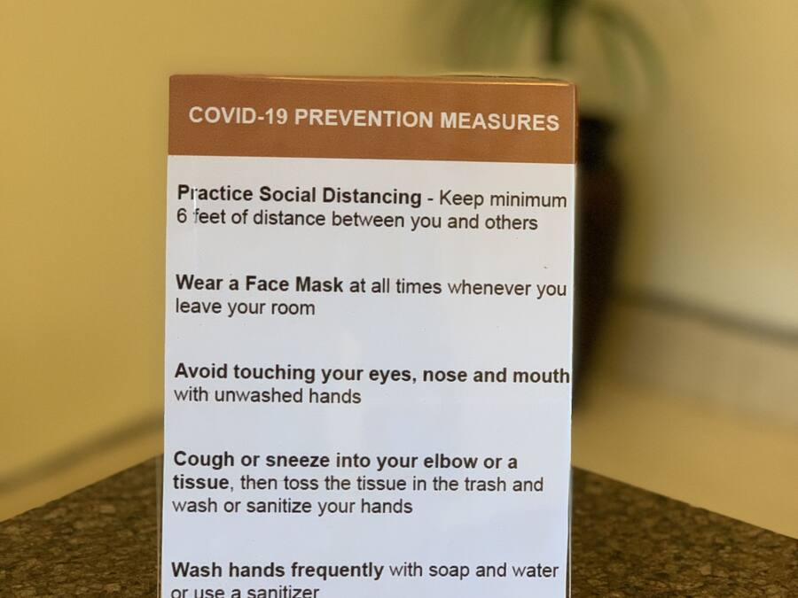A sign with COVID-19 prevention measures including social distancing, wearing a face mask, avoiding touching face, and washing hands.