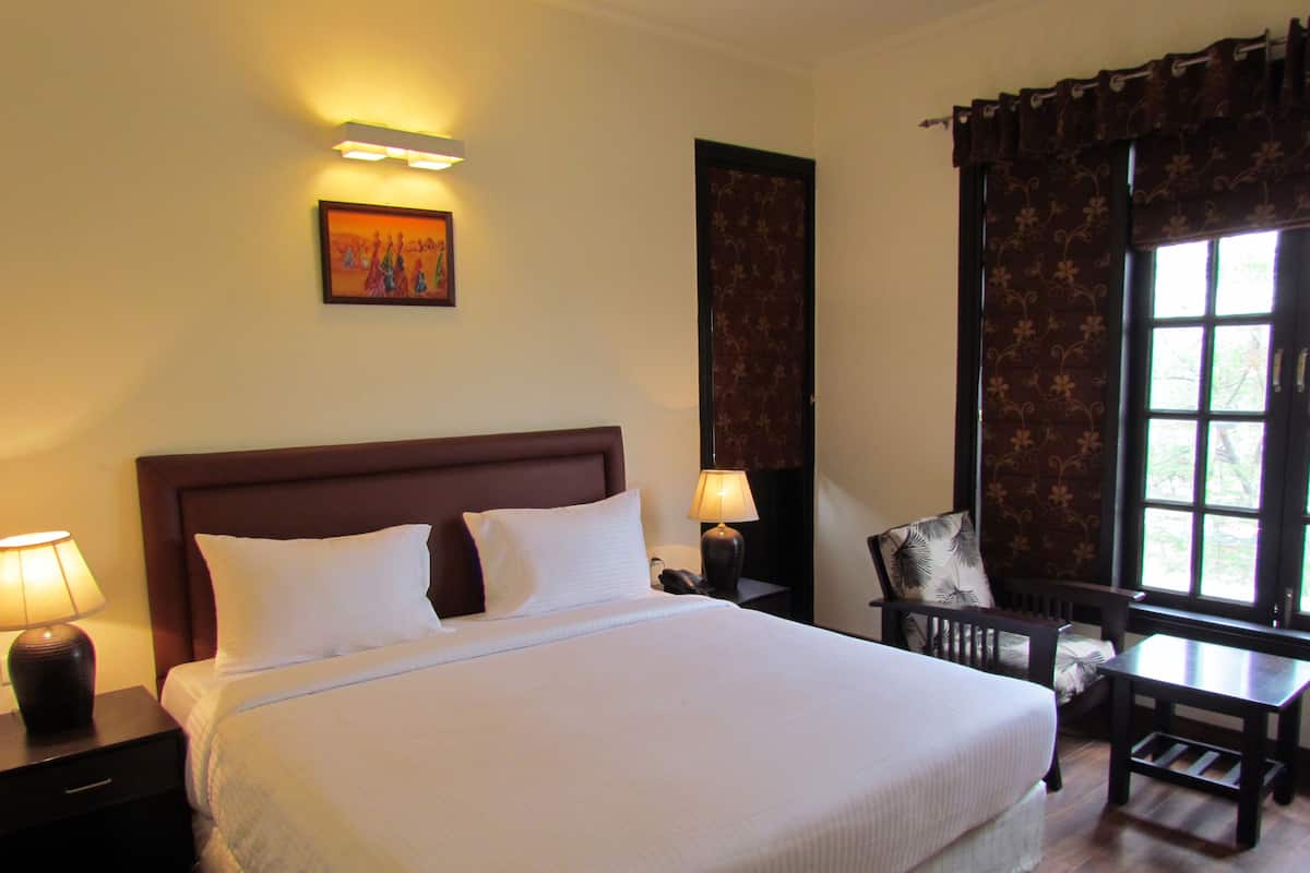 A neatly made bed with white linens, a wooden headboard, and two bedside lamps. There is a chair with a floral cushion and a small table beside the window.