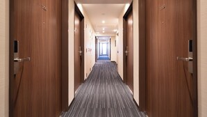 Un largo y estrecho pasillo de hotel con puertas de madera a ambos lados, que desemboca en un vestíbulo luminoso al final.