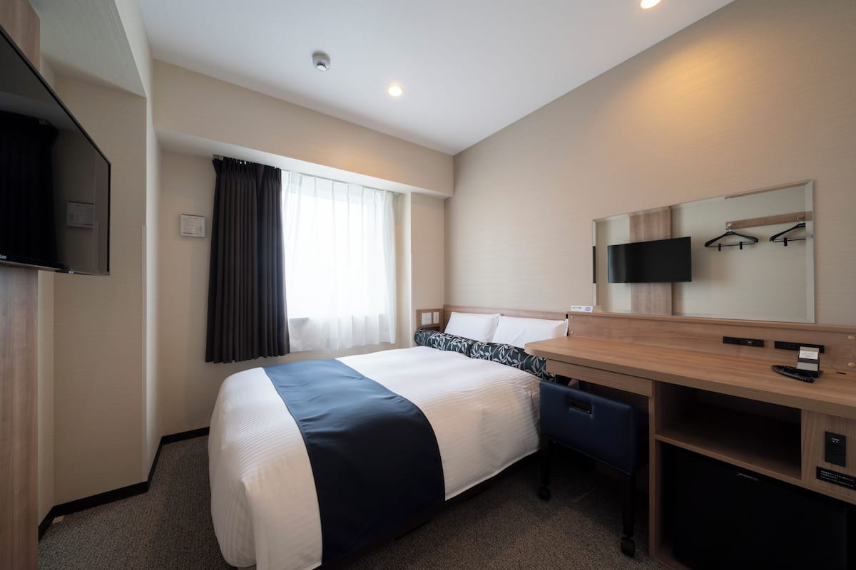Habitación de hotel con cama, escritorio, silla, televisor, espejo y ventana con cortinas.