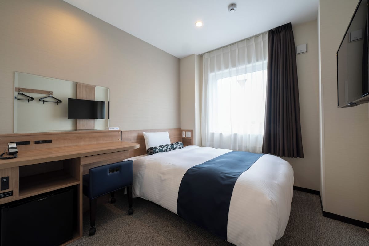 Una habitación de hotel con cama, escritorio, computadora, televisor y ventana con cortinas.